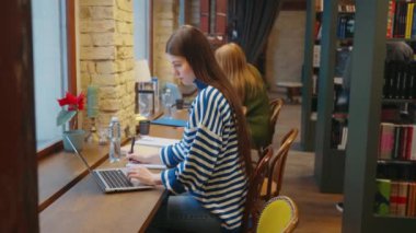 Çizgili süveter giyen bayan öğrenci kütüphanedeki ahşap masada dizüstü bilgisayarına odaklanıyor, kitaplıklar ve arka planda başka bir öğrencisiyle birlikte. Eğitim ve kaynak arama kavramı