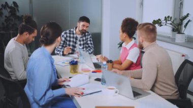 Bir grup profesyonel, parlak bir ofis yerinde bir toplantıya katıldı. Masanın etrafında dizüstü bilgisayarlar ve belgelerle ilgili tartışmalara katılan çeşitli takımlarla toplantı