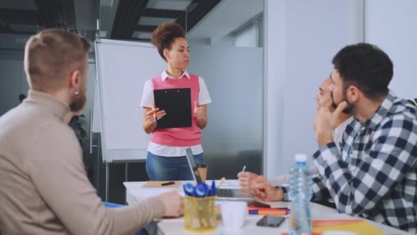 在工作会议上作报告的黑人妇女 与坐在桌旁专心工作的同事一起站在剪贴板旁 自信专业地向现代办公室的同事们介绍业务理念 — 图库视频影像