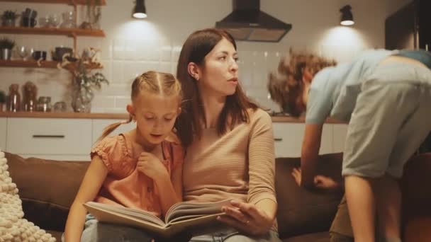 Мати Читає Своєю Дочкою Син Грайливо Стрибає Дивані Затишній Кухонній — стокове відео