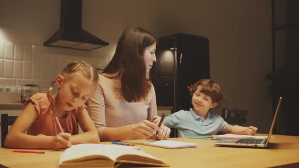 母はラップトップの仕事と娘の宿題を手伝うことの間で彼女の注意を分け 彼女の息子は満足してそれらの横に描きます — ストック動画