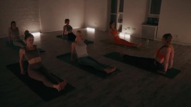 Bir grup birey yoga seansına katılıyor. Çekirdeklerine ve dengelerine odaklanıyorlar. Ters tahta duruşunu gerçekleştiriyorlar. Loş ışıklı oda akşam çalışması için sakin bir atmosfer yaratıyor.