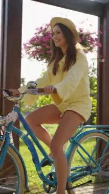Hasır şapkalı ve sarı gömlekli gülümseyen kadın mavi renkli bisiklet sürüyor, arkasında pembe çiçekler ve yeşillik var. Sıcak güneş ışığı neşeli, kaygısız sürüş atmosferini arttırır.