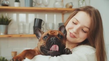 Kahverengi Fransız Bulldog 'u kucaklayan mutlu genç bir kadın. Dilini şakayla uzatan, arka planda mutfak rafları olan.