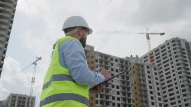 Sakallı erkek inşaat denetçisi, inşaat halindeki yüksek binaların ve vinçlerin zeminine sahip panoya yansıtıcı yelek ve yelek notları yazıyor.
