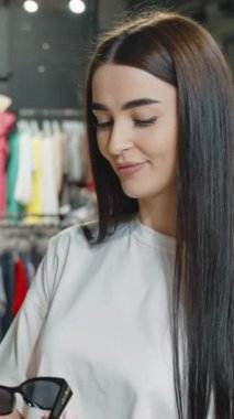 Moda mağazasında moda kıyafetleriyle çevrili şık güneş gözlüğü deneyen neşeli bir kadın. Bayan müşteri tatil için aksesuar alıyor, kameraya bakıyor ve gülümsüyor.