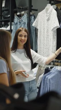 Dikey Ekran: Mağazada iki kadın elbise rafı inceliyor, biri arkadaşına göstermek için mavi gömleği seçiyor. Birlikte alışverişe çıkmıştık. Arkadaşlarıyla alışveriş yaparken gömlek seçen kadın.