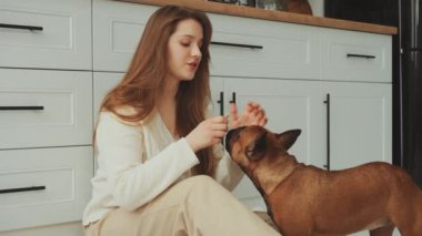 Genç bir kadın yerde Fransız Bulldog 'la oturuyor, nazikçe okşuyor. Dişi, evcil hayvanlarla sahipleri arasında özel bir bağ olan ev mutfağında köpeğiyle iç açıcı bir anın tadını çıkarır.
