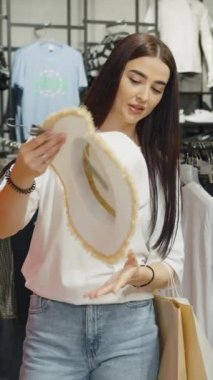 Dikey Ekran: Genç kadın arkadaşlarıyla alışveriş yaparken, arka planda çeşitli giysi rafları olan yaz şapkasını mutlulukla inceliyor. Giyim mağazasında eğlence ve moda seçimi