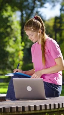 Pembe tişört ve kot pantolon giyen genç bir bayan güneşli parkta bankta oturuyor, dizüstü bilgisayar kullanıyor ve egzersiz kitabına yazıyor, saçlarını savuruyor. Öğrenci dışarıda çalışıyor. Çevrimiçi eğitim kavramı