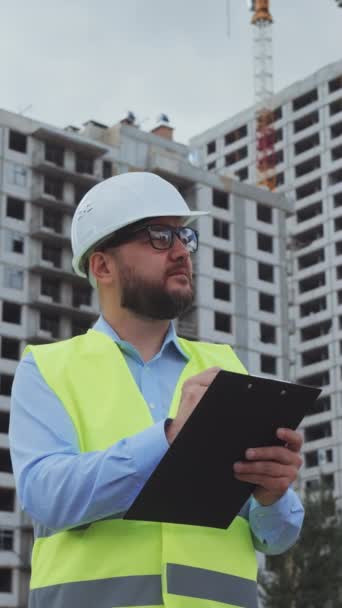 垂直屏风 带胡子的建筑检查员 戴着硬礼帽 身穿安全背心 在纸上做笔记 背景是多层房屋 低角度人员检查建筑工地 控制的概念 — 图库视频影像
