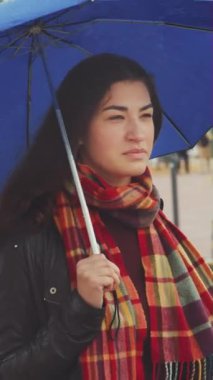 Kareli atkı takan çekici genç bir kadın, yağmur altında cadde kavşağının önünde mavi bir şemsiyeyle bekliyor. Arabalar ve arka plandaki insanlar bulanık. Şehir hayatı kavramı
