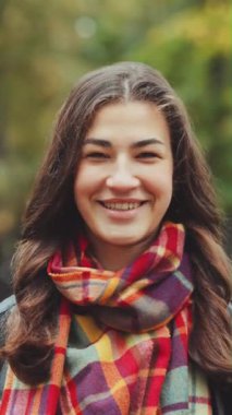 Dikey Ekran: Uzun saçlı, deri ceketli, kareli atkısı olan genç bir kadın rüzgarlı sonbahar parkında kameraya gülümsüyor. Mutlu insan portresi. Duygu kavramı