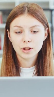 Dikey Ekran: Konsantre genç kadın video konferansına katılıyor, kitaplığa karşı dizüstü bilgisayara odaklanıyor