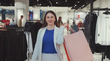 Kadın müşteri başarılı bir eğlence sonrası alışveriş torbaları taşıyor, kameraya gülümsüyor. Mutlu kadın, çağdaş moda mağazasında başarılı alışverişlerinin keyfini çıkarıyor.