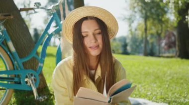 Saman şapkalı genç kadın kitap okumaya dalmış, arka planda mavi bisikletle güneşli parkta çimlere oturmuş.