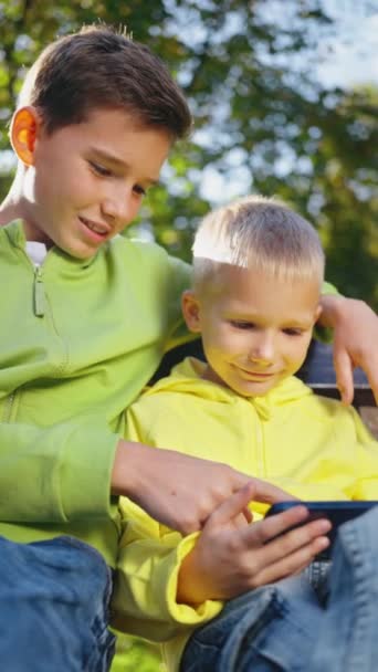 垂直屏幕 两个兄弟为孩子玩电子游戏 分享数字小玩艺儿 兄弟姐妹们在公园的长椅上放松 在手机上使用在线父母控制应用程序 移动上网成瘾 — 图库视频影像