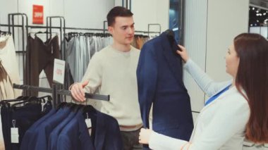 Erkek alışverişçi kadın tezgahtardan yardım alırken, iyi organize edilmiş giyim mağazasından klasik mavi takımı seçiyor.
