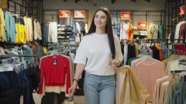 Elinde alışveriş torbalarıyla keyifli genç bir kadın etrafındaki çeşitli kıyafetlerle moda mağazasına doğru yürüyor, memnuniyet ve başarılı perakende terapisi ile.