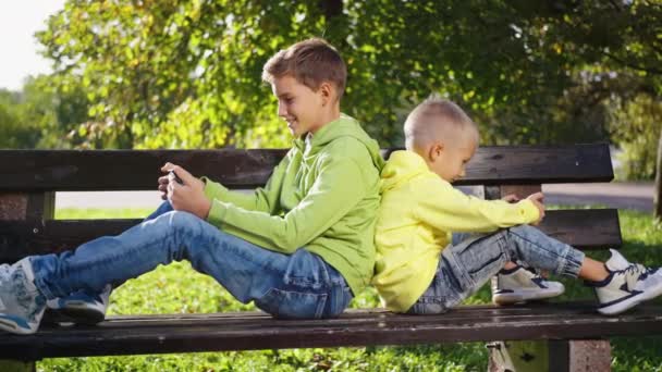 兄弟俩都沉浸在自己的数码世界里 背靠背地坐在公园的长椅上 带着他们的小玩意在户外享受着阳光灿烂的一天 — 图库视频影像