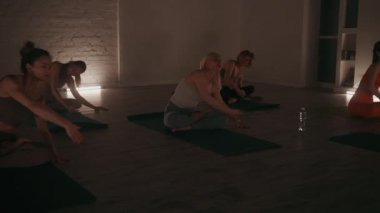 Yoga kursuna katılan bir grup kadın, sıcak ışıklandırılmış, tuğla duvarlı bir stüdyoda paspaspaslarının üzerine uzanarak esneklik ve gevşemeyi teşvik ediyor.