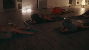 Yoga seansına katılanlar rahat, loş ışıklandırmalı stüdyoda paspaslar üzerinde poz vererek destek topluluğunda rahatlama ve esneklik gösteriyorlar. Kadınlar derin esneme ve farkındalık pratikleriyle meşgul olurlar.