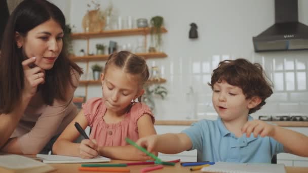 母親は 子供の教育における両親の役割を育成するキッチンテーブルで宿題を持つ彼女の2人の幼い子供を注意深く助けます — ストック動画