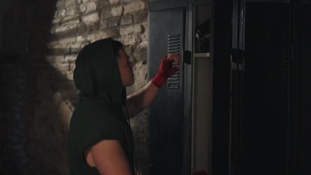 緑のフーディーの男性ボクサーは 素朴なレンガの壁で薄暗く点灯ロッカールームで彼のボクシング手袋をぶら下げ トレーニングのために準備します — ストック動画