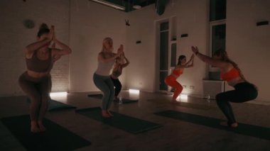 Yoga sınıfına katılan bir grup kadın, hafif ışıklandırılmış stüdyoda kompleks pozlar vererek, konsantrasyon, denge ve huzurlu çalışma ortamı üzerine odaklanırlar.