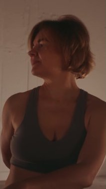 Dikey ekran: Kadın loş aydınlatılmış odada yoga çevirme alıştırması yapıyor, sükuneti ve farkındalığı yansıtıyor. Sağlık ve grup fitness konsepti