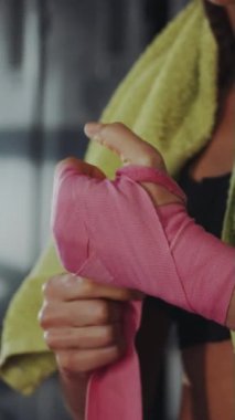 Dikey ekran: Ellerine pembe boks örtüsü saran, omzuna havlu sarılı bir kadın, zorlu spor salonundaki antrenman oturumuna hazırlanıyor