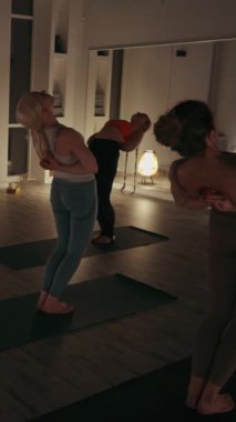 Dikey ekran: Yoga sınıfındaki kadınlar loş ışıklı odada esneme hareketleri yapıyorlar. Bir grup birey, yumuşak ışıklandırmayla aydınlatılmış akşam stüdyosunda ters köşe pozları veriyor.