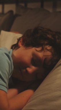 Dikey ekran: Mavi pijama giyen genç çocuk huzursuzca yatakta döner, oyuncak ayısı yanında yatarken uyumaya çalışır.