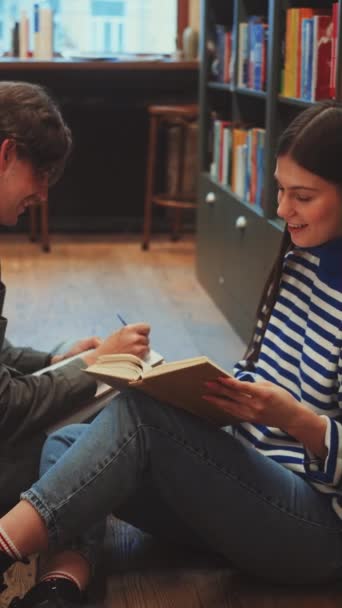 垂直屏幕 男女学生坐在图书馆过道的地板上 全神贯注地阅读和讨论书籍 迷失在共同发现和安静的友谊中 — 图库视频影像