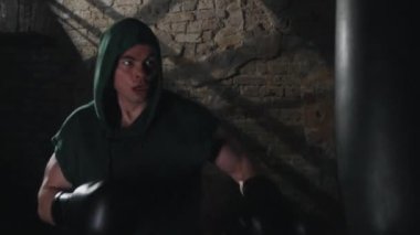 Koyu yeşil kapüşonlu ve siyah eldivenli erkek boksör, eski tuğla duvarlı loş ışıklı spor salonunda ağır çantada pratik yapıyor. Boks eğitimine adanmışlık ve sıkı çalışma