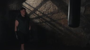 Yeşil tişörtlü konsantre boksör, düşünceli ve bitkin görünüyor, antrenman sırasında loş ışıklı spor salonunda kırsal tuğla zemin ve yakınındaki kum torbasıyla