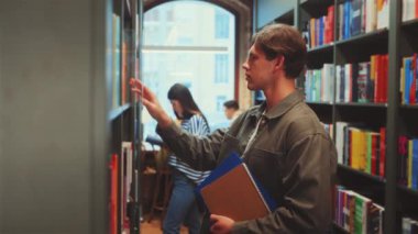 Adam rafları karıştırır ve modern kütüphanede kitap seçer, elinde defterler, diğer insanlarla birlikte arka planda çalışır, renkli kitaplarla çevrili, eğitici aktivitelerle meşgul..