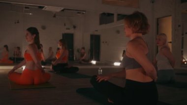Çağdaş stüdyo ortamında, aynalar ve sıcak ışıklandırma ile yoga yapan, meditasyon ve akşam dersleri sırasında duruş üzerine odaklanan dingin bir grup kadın.