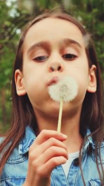 Dikey ekran: kot ceketli genç kız karahindibaya üfleyerek orman parkında tohumları uçuruyor. Neşeli bir an, doğanın ve çocukluğun basit eğlenceleri.