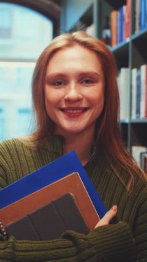 Dikey ekran: Gülümseyen öğrenci kütüphanedeki kitaplıklar ve diğer müşteriler kitaplara bakarken elinde defterler ve tablet tutuyor. Öğrenim ve öğrenim atmosferi