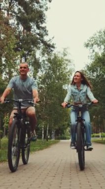 Gülümseyen çiftler park yolu boyunca bisiklet sürerler. Erkek ve kadın, yemyeşil ve ağaçlarla çevrili bisiklet sürerek neşeli bir anı paylaşıyor. Açık hava aktivitesi sağlıklı yaşam tarzını teşvik eder
