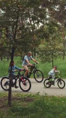 Dikey ekran: Baba ve iki çocuk bir yaz günü manzaralı bir parkta bisiklet sürmekten zevk alıyorlar. Aile zamanı, açık hava aktiviteleri ve sağlıklı yaşam tarzı kavramı.