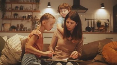 Anne, sıcak, rahat bir mutfak ortamında iki çocukla kitap okuyor, öğrenme ve kaynaşmayı teşvik ediyor. Oğlumuz annesinin omzuna yaslanıyor. Aile, eğitim ve birliktelik kavramı.