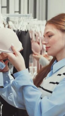 Dikey ekran: Genç kadın giyim mağazasında sutyen inceliyor, diğer iç çamaşırı seçenekleri arasında stil ve kalitesini dikkatlice değerlendiriyor