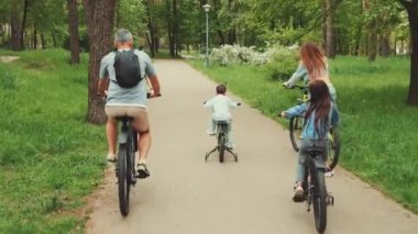 Yeşillik bir parkta, asfalt bir yolda birlikte bisiklete binen aile. Ebeveynler ve çocuklar doğa aktivitelerinin keyfini çıkarıyorlar. Aile bağı ve sağlıklı yaşam tarzı kavramı