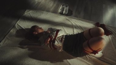 Halıda uzanmış kırmızı iplerle Shibari sanatını icra eden bir kadın. Dizginleme ve incinebilirlik teması. Beyaz spor ayakkabıları bir kenara bırakıldı. Sanatsal ifade ve gelenek kavramı