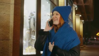 Mavi kışlık giyinmiş bir kadın telefonda konuşuyor, gülümsüyor, geceleri arka planda şehir ışıklarıyla dükkanın önünde dikiliyor. Sıcak, neşeli bir atmosfer. Kış kavramı, iletişim ve neşe