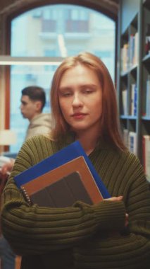 Dikey ekran: Uzun kızıl saçlı, elinde defterler olan düşünceli genç kadın, rahat bir kütüphane ortamında renkli kitaplıkların arasında duruyor. Eğitim ve öğrenme kavramı