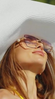 Güneş gözlüklü ve geniş kenarlı şapkalı bir kadın yazın havuzda dinleniyor. Zarafet, tarz ve rahatlamaya odaklan. Şık aksesuarlarla güneşli bir günün tadını çıkarma kavramı