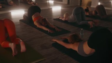 Uzatılmış yavru köpek pozu veren bir grup kadın loş ışıklı stüdyoda yoga minderlerinde poz veriyor. Sıcak ortam ışıkları ve aynalar huzurlu atmosferi geliştirir. Rahatlama ve esneklik kavramı